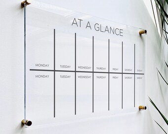 May include: A clear acrylic weekly planner with the text "AT A GLANCE" at the top. The planner has seven columns for each day of the week, with the days of the week labeled at the top and bottom.
