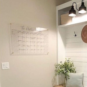 May include: A white entryway bench with a wooden seat and a white shelf above. The shelf has a woven basket and a straw hat. There are two hooks on the wall above the shelf. The floor is black and white herringbone tile. The text "The Cheston Family" is visible on a clear acrylic calendar hanging on the wall.