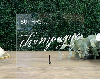 May include: Clear acrylic sign with the text "But first... champagne" in white cursive font. The sign is sitting on a wooden surface.