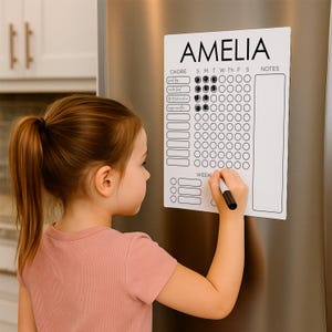 May include: A white chore chart with the name "AMELIA" at the top. The chart has columns for days of the week and rows for chores. A young girl is using a black marker to fill in the chart.