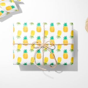 May include: A wrapped gift with pineapple-patterned paper, tied with twine. The paper features yellow pineapples with green tops on a white background. Scissors and a roll of matching paper are nearby.