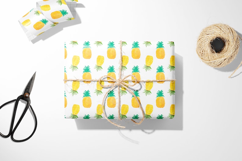 May include: A wrapped gift with pineapple-patterned paper, tied with twine. The paper features yellow pineapples with green tops on a white background. Scissors and a roll of matching paper are nearby.
