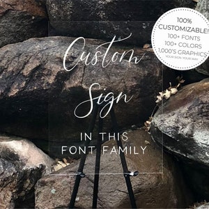 May include: A clear acrylic sign with the text "Custom Sign" in a cursive font. The sign also includes the text "IN THIS FONT FAMILY" and a small circle with the text "100% CUSTOMIZABLE! 100+ FONTS 100+ COLORS 1,000'S GRAPHICS YOUR SIGN YOUR WAY". The sign is standing on a black tripod.