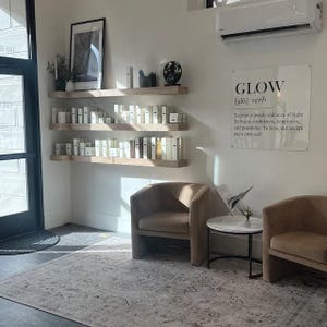 May include: A white wall with a wooden shelf displaying rows of small bottles. Two brown chairs are in front of the shelf, and a white table with a silver object on top sits between them. A sign on the wall reads "GLOW [glo] verb To emit a steady radiance of light. To beam confidence, happiness, and positivity. To love and accept one's own self."