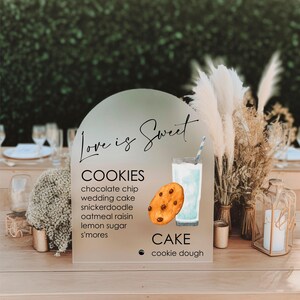 May include: A white sign with an arch shape at the top. The sign says "Love is Sweet" in cursive writing. Below the text, there is a list of cookies: chocolate chip, wedding cake, snickerdoodle, oatmeal raisin, lemon sugar, and s'mores. Below the list of cookies, there is a list of cake: cookie dough. The sign is on a wooden table with a glass of milk and a cookie next to it.