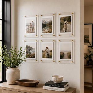 May include: A set of six clear acrylic wall-mounted photo frames arranged in two rows of three. Each frame features gold-toned hardware at the corners to secure the prints. The frames display various outdoor landscape and family portrait photographs.