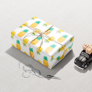 May include: A wrapped gift with a pineapple pattern, tied with twine, sits next to a black toy car carrying small packages. Decorative silver scissors are also present on a light gray surface. The scene suggests a gift-giving or travel theme.