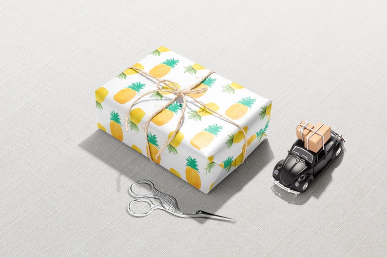 May include: A wrapped gift with a pineapple pattern, tied with twine, sits next to a black toy car carrying small packages. Decorative silver scissors are also present on a light gray surface. The scene suggests a gift-giving or travel theme.