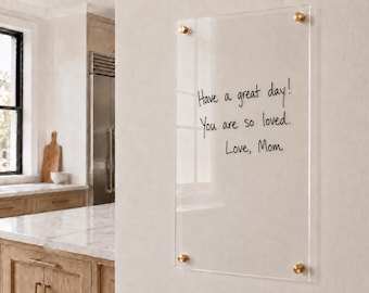 May include: A rectangular clear acrylic wall-mounted dry erase board secured with four gold-toned metal standoffs. The surface features black handwritten text that reads: Have a great day! You are so loved. Love, Mom.
