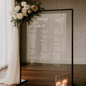 May include: Clear acrylic wedding seating chart with a black metal frame, adorned with a floral arrangement of white roses and greenery. The chart displays guest names and table assignments. Candles in glass cylinders are at the base.