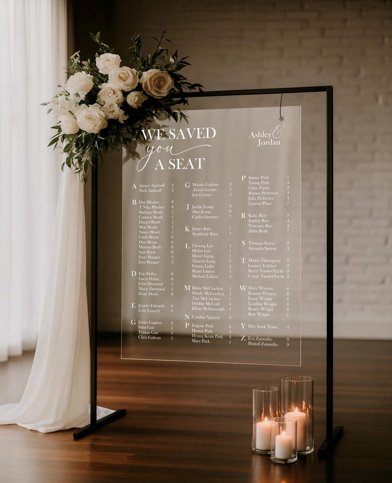 May include: Clear acrylic wedding seating chart with a black metal frame, adorned with a floral arrangement of white roses and greenery. The chart displays guest names and table assignments. Candles in glass cylinders are at the base.