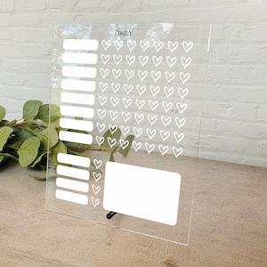 May include: A clear acrylic daily planner with a grid of white hearts and blank spaces for writing. The planner has the word "DAILY" printed at the top.