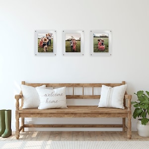 May include: Three clear acrylic photo frames with black mounting hardware, each displaying a color photograph of a family in a field. The frames are arranged horizontally on a white wall above a wooden bench with two white pillows and a green plant.