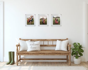 May include: Three clear acrylic photo frames with black mounting hardware, each displaying a color photograph of a family in a field. The frames are arranged horizontally on a white wall above a wooden bench with two white pillows and a green plant.