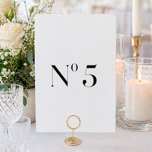 May include: A white table number card with the number "N°5" in black, held by a gold wire holder. The card is on a white tablecloth, with flowers, candles, and glassware in the background.