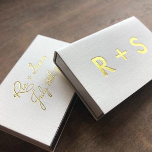 May include: Two white boxes with gold lettering. The top box says "R+S" and the bottom box says "Ria + Sean July 14th, 2023".
