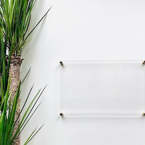 May include: A clear acrylic frame with gold metal corners, hanging on a white wall. The frame is empty and ready for a photo or artwork.