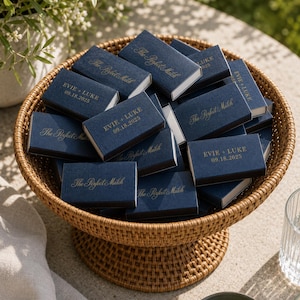 May include: A collection of rectangular navy blue matchboxes with gold foil text. Some boxes feature the phrase The Perfect Match, while others are personalized with the names EVIE + LUKE and the date 09.18.2025, all arranged in a woven basket.
