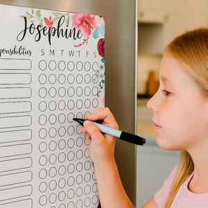 May include: A white magnetic chore chart with floral accents and the name "Josephine" in cursive. The chart includes spaces for responsibilities and a weekly tracker. A person is using a black marker to fill in the chart.