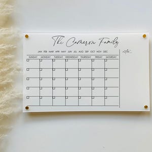 May include: White acrylic calendar with gold push pins. The calendar has a black header that reads "The Carmeron Family". The calendar has a month and day of the week header and a grid of squares for each day of the month.