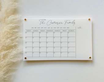 May include: White acrylic calendar with gold push pins. The calendar has a black header that reads "The Carmeron Family". The calendar has a month and day of the week header and a grid of squares for each day of the month.