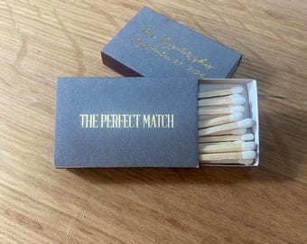 May include: A gray matchbox with the text "The Perfect Match" printed in gold on the front. The matchbox is open and contains a stack of wooden matches with white tips.