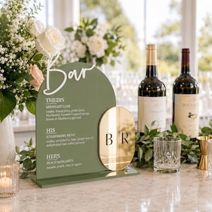 May include: An arched olive green acrylic bar menu sign with white text reading Bar, Theirs, His, and Hers, featuring a gold oval mirror accent with the letters B and R. The sign lists cocktail ingredients and sits on a rectangular base.