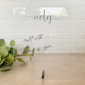 May include: A clear acrylic notes board with the word "notes" in cursive script at the top. Below, the text "call with Thomas @ 3pm" is written. A black pen rests at the bottom. Green foliage is visible on the left.