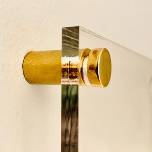 May include: A close-up of a brass screw and stand used to mount a clear acrylic plaque to a wall.