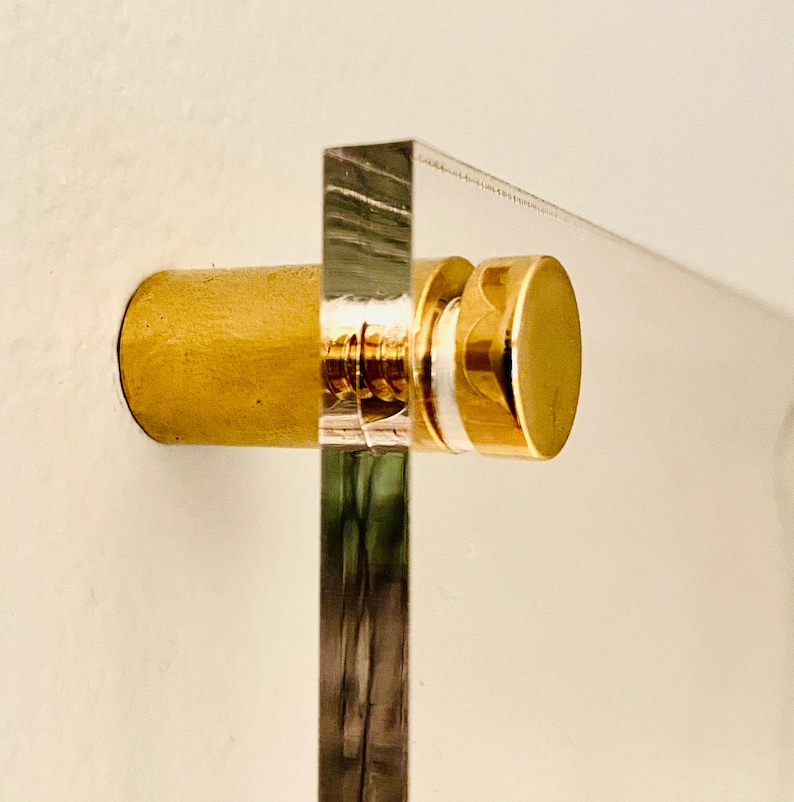 May include: A close-up of a brass screw and stand used to mount a clear acrylic plaque to a wall.