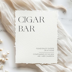 May include: A white rectangular sign with the words "CIGAR BAR" in black, elegant font. Below, smaller text reads "PLEASE ENJOY OUTSIDE OR AT HOME" and "COMPLIMENTS OF THE NEW MR + MRS CAMERON". The sign has a textured paper appearance.