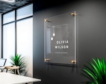 May include: A clear acrylic sign with gold standoffs, featuring a white logo with two hanging light bulbs and the text "Olivia Wilson Interior Designer".
