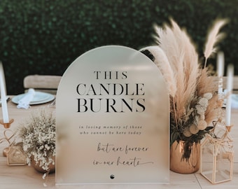 May include: A white acrylic sign with a rounded arch top reads "THIS CANDLE BURNS in loving memory of those who cannot be here today but are forever in our hearts". The sign is on a wooden table with a vase of white flowers and a gold lantern.