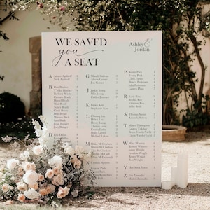 May include: A wedding seating chart on a white sign with the text "WE SAVED YOU A SEAT" in elegant script. The names of the guests are listed alphabetically. The sign is decorated with a floral arrangement in soft colors.