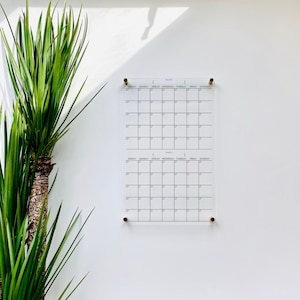 May include: Two blank monthly calendars on a white wall. The calendars are printed on clear acrylic and have a grid layout with days of the week labeled. The calendars are hung with gold push pins.