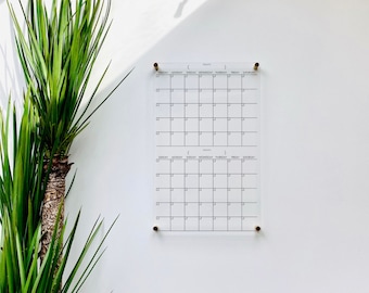 May include: Two blank monthly calendars on a white wall. The calendars are printed on clear acrylic and have a grid layout with days of the week labelled. The calendars are hung with gold push pins.
