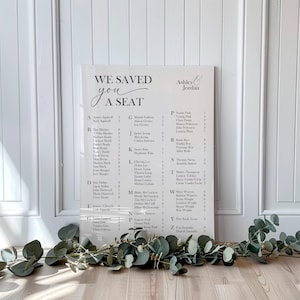 May include: A white wedding seating chart with the text "WE SAVED YOU A SEAT" and the names of guests. The names are arranged alphabetically with the bride and groom's names at the top. Eucalyptus greenery decorates the bottom.