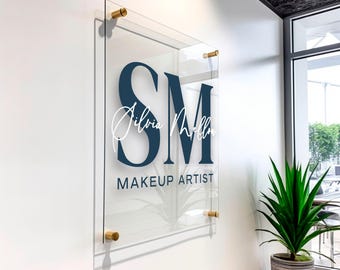Può includere: Un cartello in vetro trasparente con un logo blu e bianco. Il logo è composto dalle lettere "SM" in grassetto. Il testo "Silvia Mellon" è scritto in corsivo sotto le lettere. Il testo "MAKEUP ARTIST" è scritto sotto il nome.
