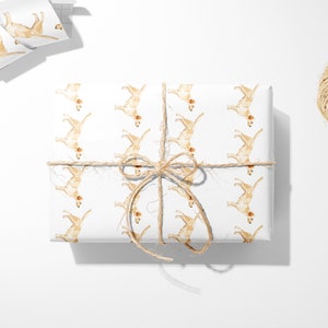 May include: White gift wrap featuring a repeating pattern of watercolor-style golden retriever illustrations. The wrapped package is tied with twine. A roll of matching wrapping paper, scissors, and a ball of twine are also visible.