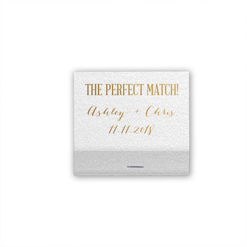May include: A white matchbox with gold text that reads "THE PERFECT MATCH!" followed by "Ashley + Chris" and the date "11.11.2018". The bottom portion of the matchbox is a light gray color.
