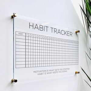 May include: A clear acrylic habit tracker with a grid for marking off daily habits. The tracker has the text "HABIT TRACKER" at the top and the quote "MOTIVATION IS WHAT GETS YOU STARTED. HABIT IS WHAT KEEPS YOU GOING." at the bottom.