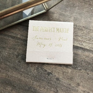 May include: A small, off-white matchbox with gold lettering that reads "THE PERFECT MATCH!" followed by names and a date. A gold-colored picture frame is in the background, resting on a wooden surface.