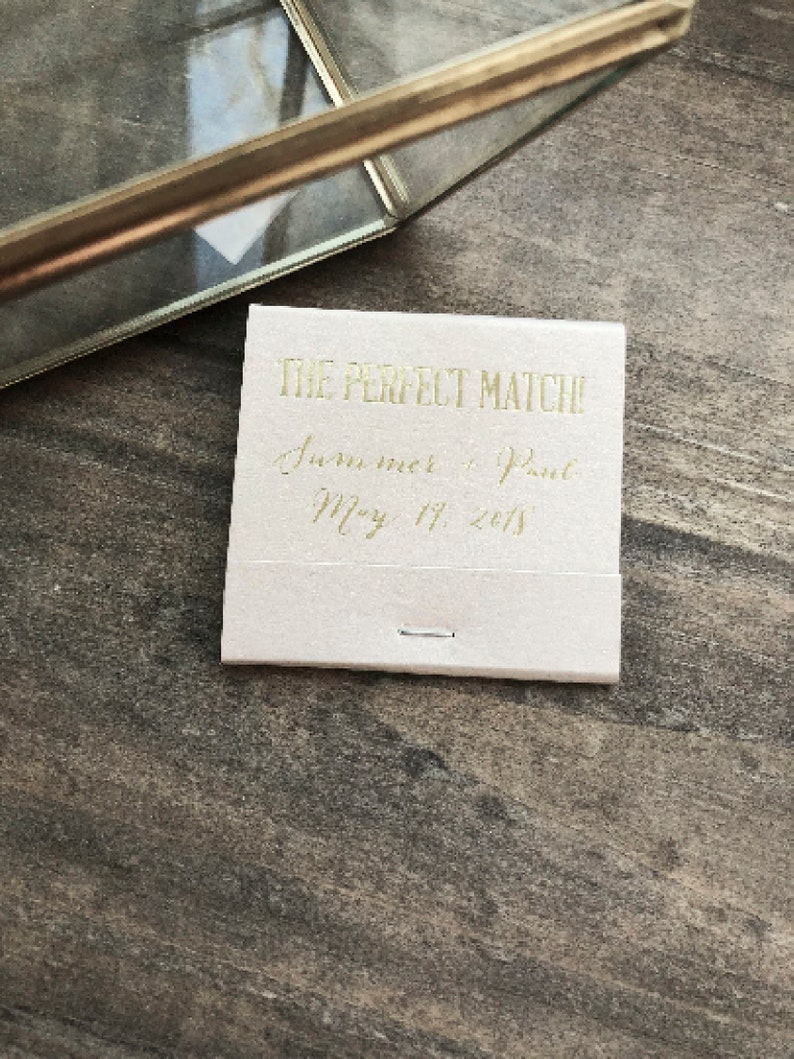 May include: A small, off-white matchbox with gold lettering that reads "THE PERFECT MATCH!" followed by names and a date. A gold-colored picture frame is in the background, resting on a wooden surface.