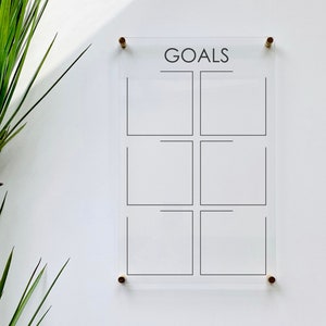 May include: A clear acrylic board with the word "GOALS" printed in black at the top. The board is divided into six sections with black lines, creating a grid for writing down goals.