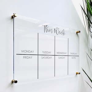 May include: A clear acrylic weekly planner with gold metal hanging hardware. The planner has spaces for each day of the week, Monday through Sunday, and a notes section. The text "This Week" is printed at the top of the planner.