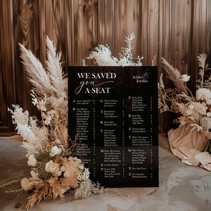 May include: A black wedding seating chart with the text "WE SAVED YOU A SEAT" and names listed alphabetically. The names are in white font. The sign is decorated with dried flowers and pampas grass.