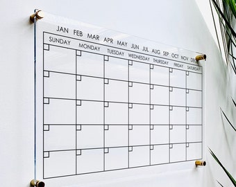 May include: A clear acrylic monthly calendar with black grid lines and text. The calendar is mounted on a white wall with brass-colored hardware. The months of the year and days of the week are printed at the top and side.