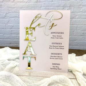 May include: A pink acrylic sign with gold lettering that reads "Let's Eat". The sign lists appetizers, entrees, desserts, and drinks.