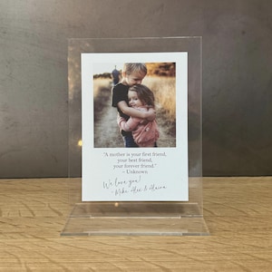 May include: A clear acrylic photo frame with a white mat and a photo of two children hugging. The photo is accompanied by a quote: "A mother is your first friend, your best friend, your forever friend." - Unknown. The frame also includes a handwritten message: "We love you! - Mike, Alex & Alaina."