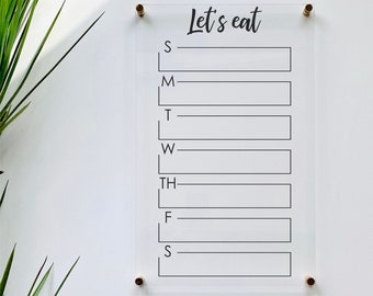 May include: Clear acrylic weekly meal planner with the words "Let's eat" in black script at the top. The days of the week are listed below with rectangular boxes for each day. The planner is mounted with gold-colored hardware.
